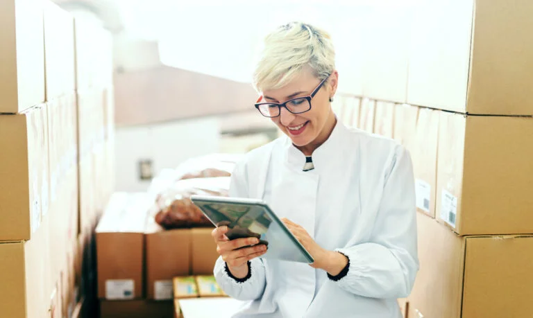 Happy Caucasian blonde employee suing tablet for work while sitting around boxes in food factory.